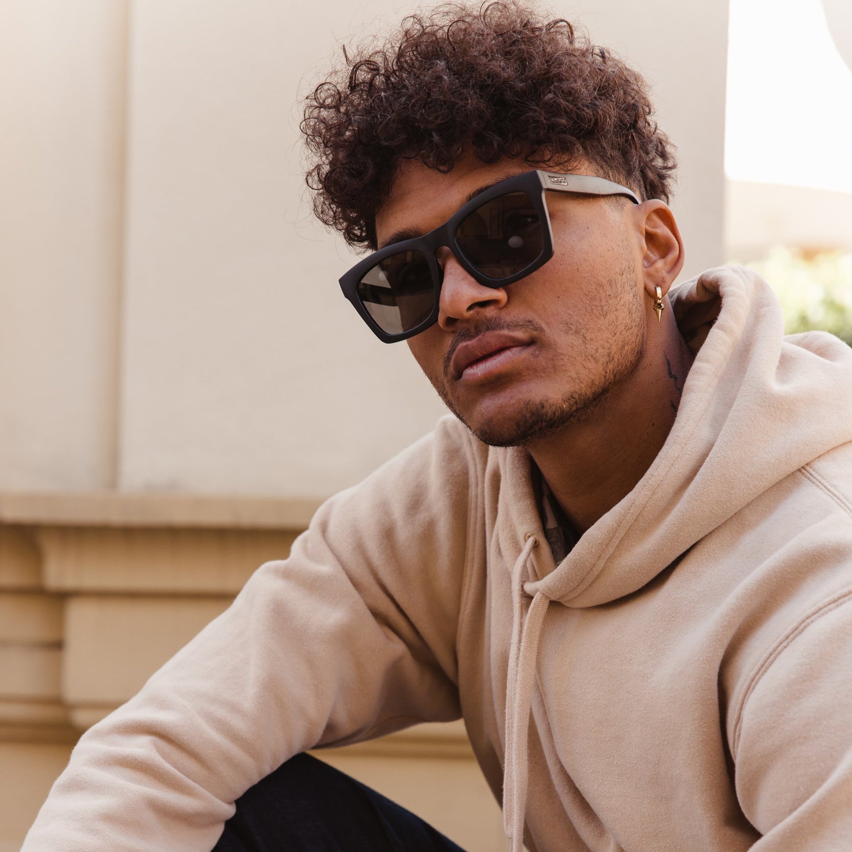 Man wearing a beige hoodie and sunglasses sitting outdoors.