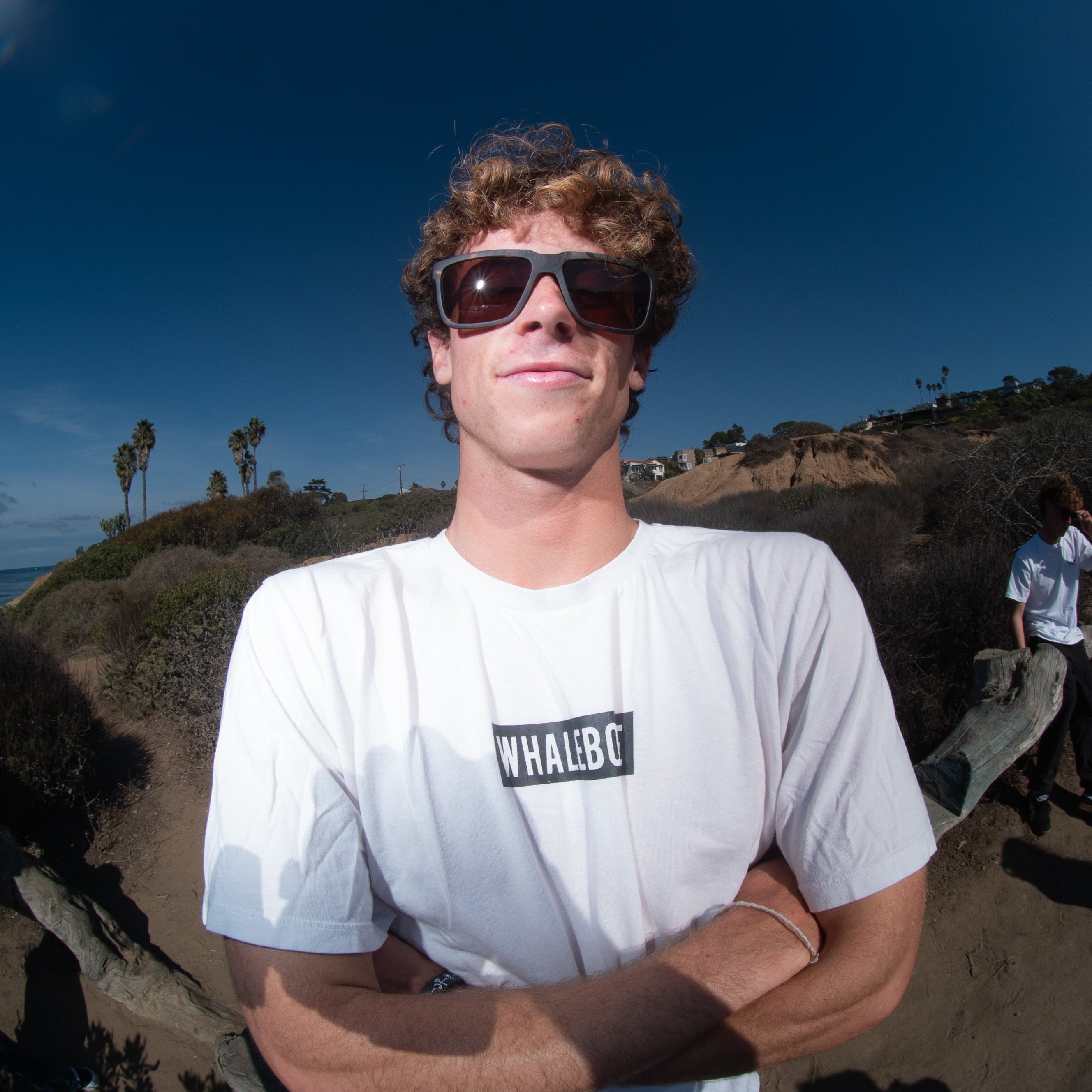 Person wearing sunglasses and a white t-shirt with a logo, standing on a coastal path.
