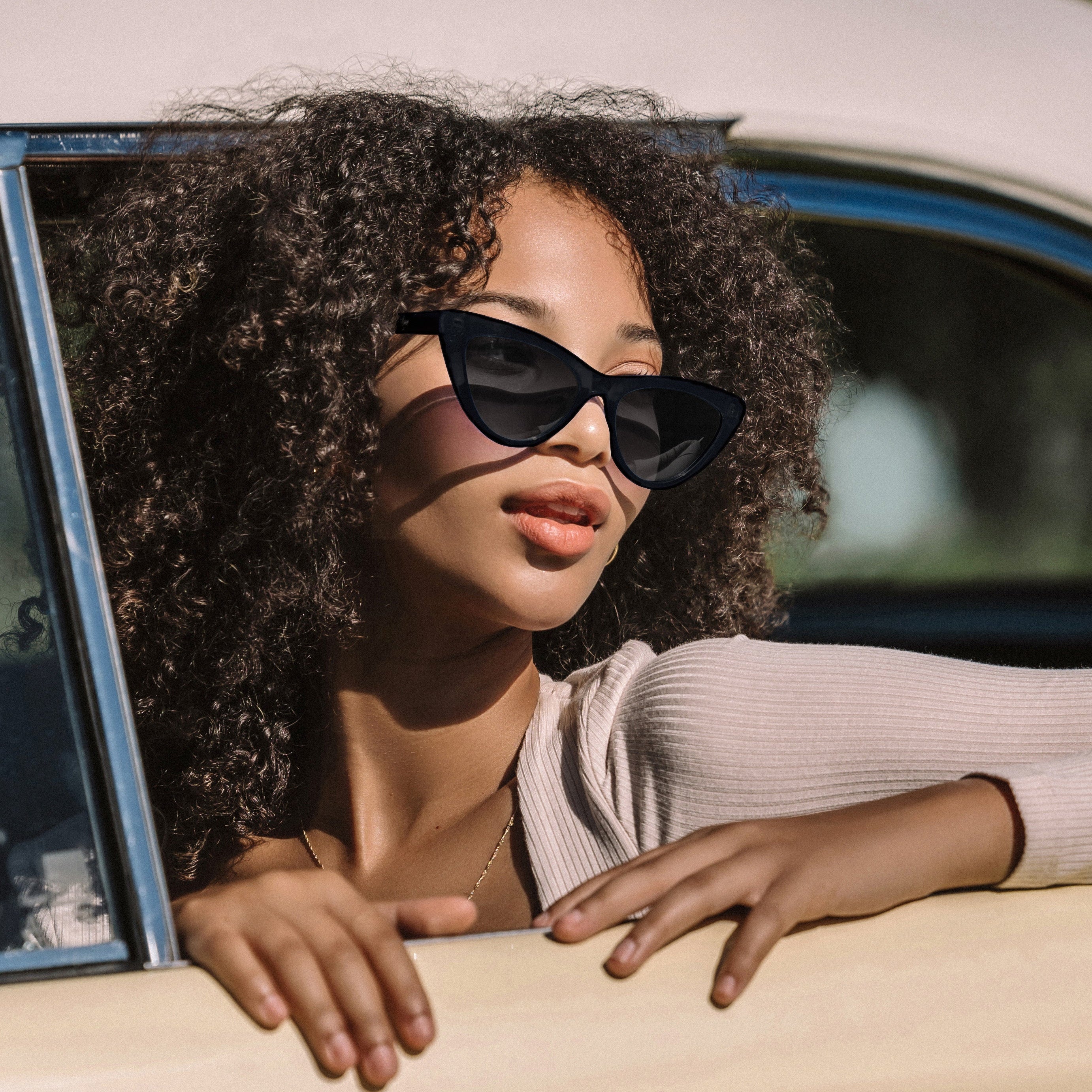 Woman wearing sunglasses and a beige top, leaning out of a car window.