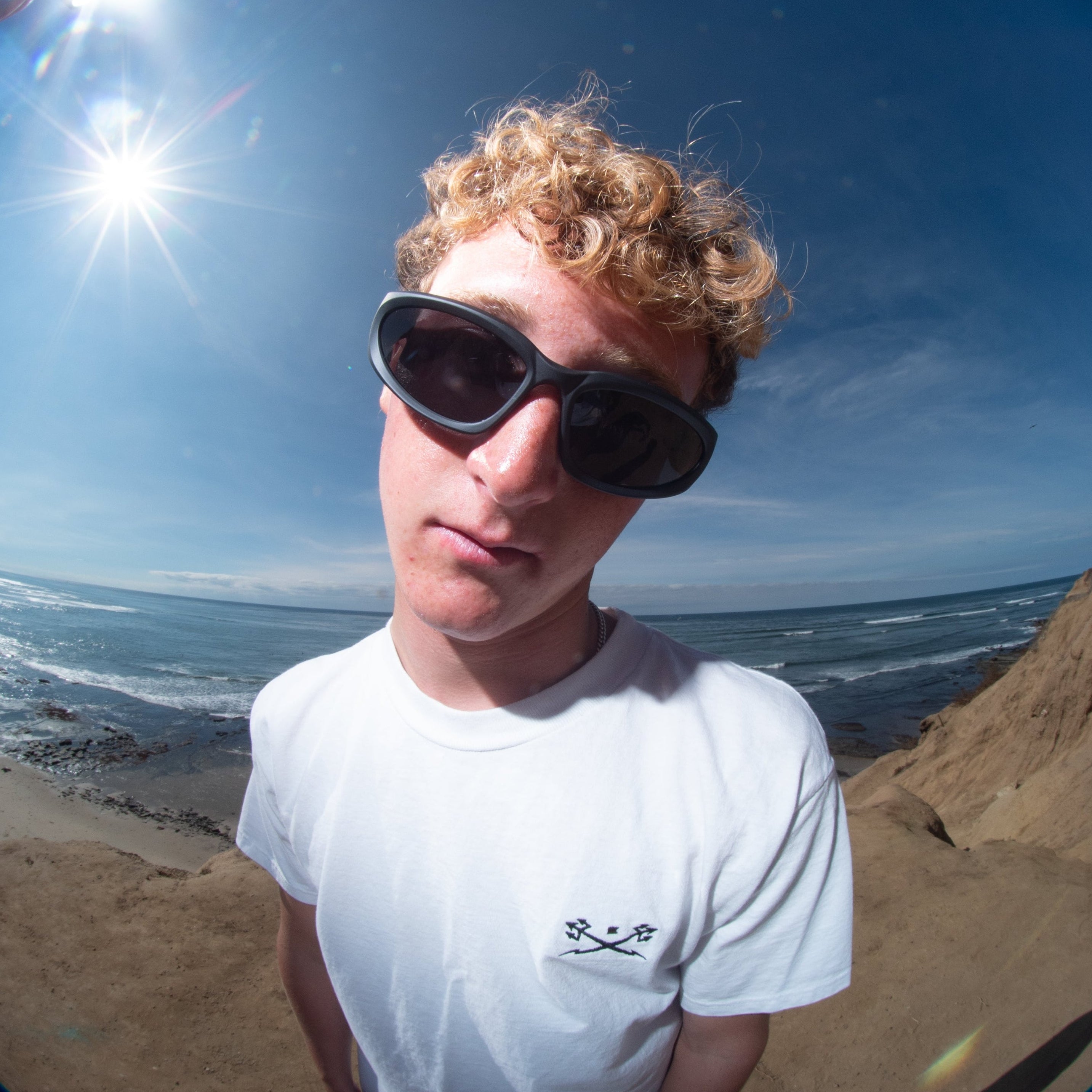 Person wearing sunglasses and a white t-shirt on a beach with a fish-eye lens effect.
