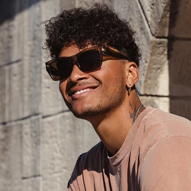Man wearing sunglasses and a brown shirt against a stone wall