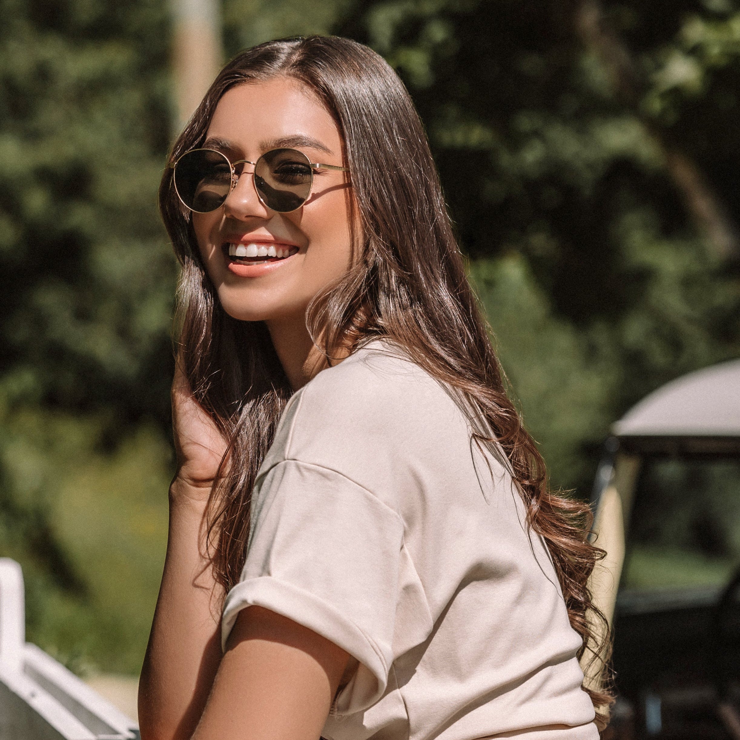 Woman in denim shorts and sunglasses leaning against a white picket fence with a vintage car in the background.