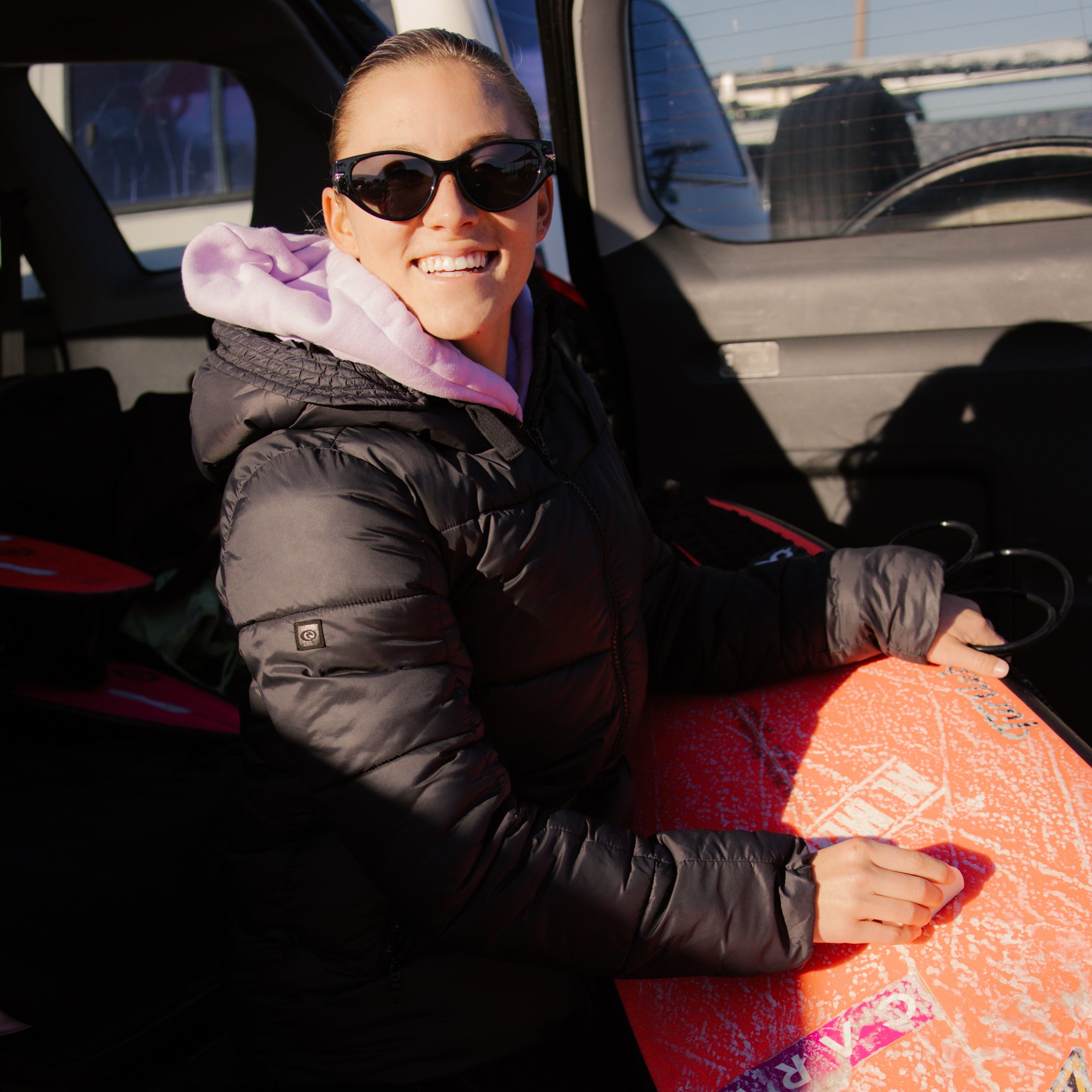 Person holding a pink surfboard, sitting in a vehicle.