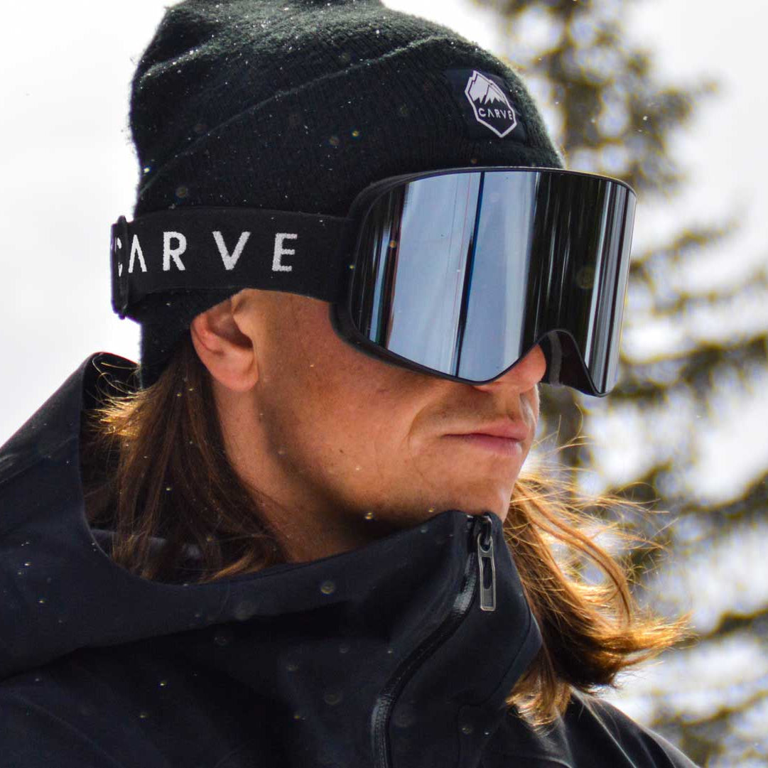 Person wearing a black beanie and goggles with 'Carve' branding, standing against a snowy background.