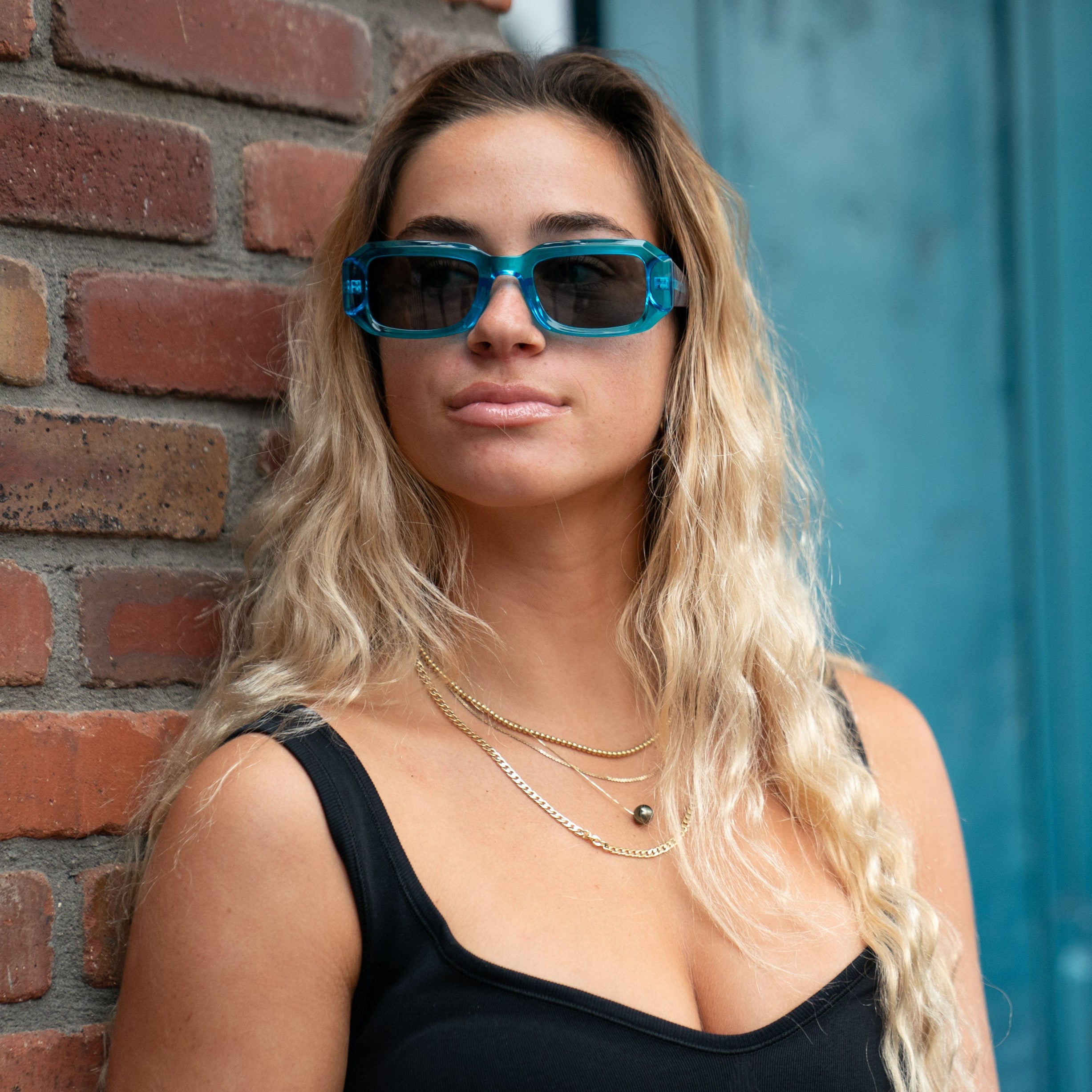 Woman wearing blue sunglasses standing against a brick wall with a blue door in the background