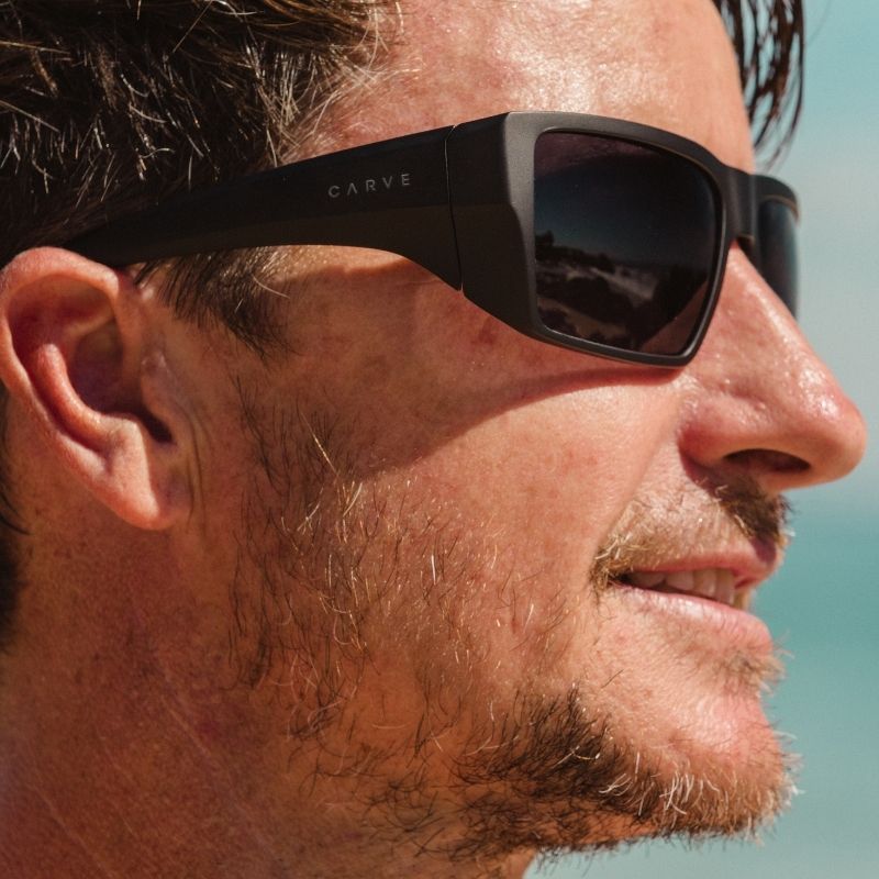 Close-up of a person wearing black sunglasses with 'Carve' branding, against a clear blue sky.