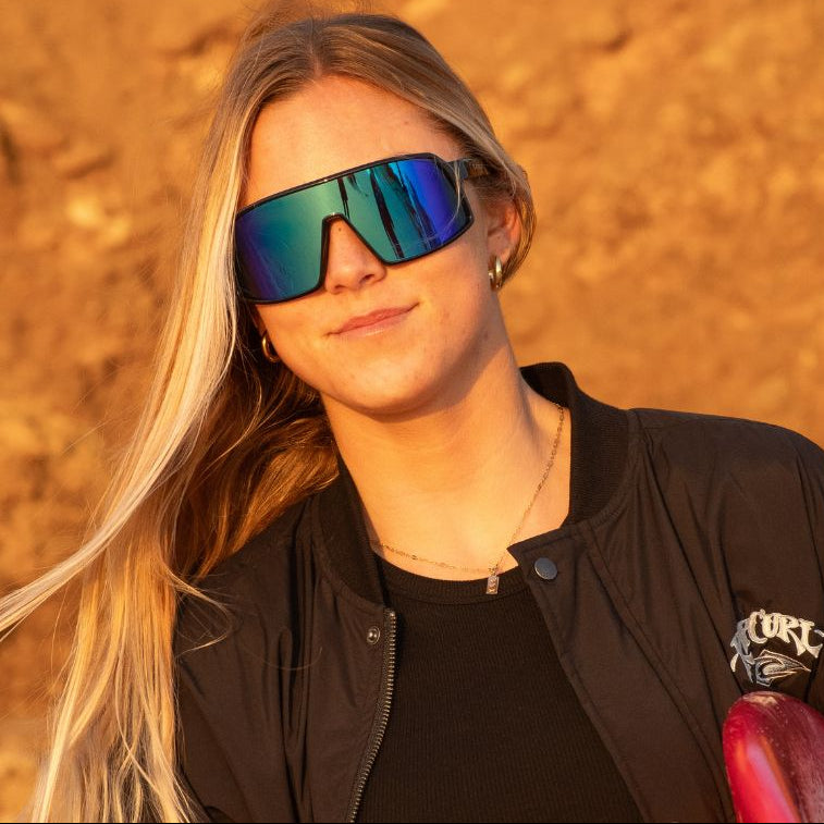 Person wearing reflective sunglasses and a black jacket with a logo, standing against an orange-brown background.