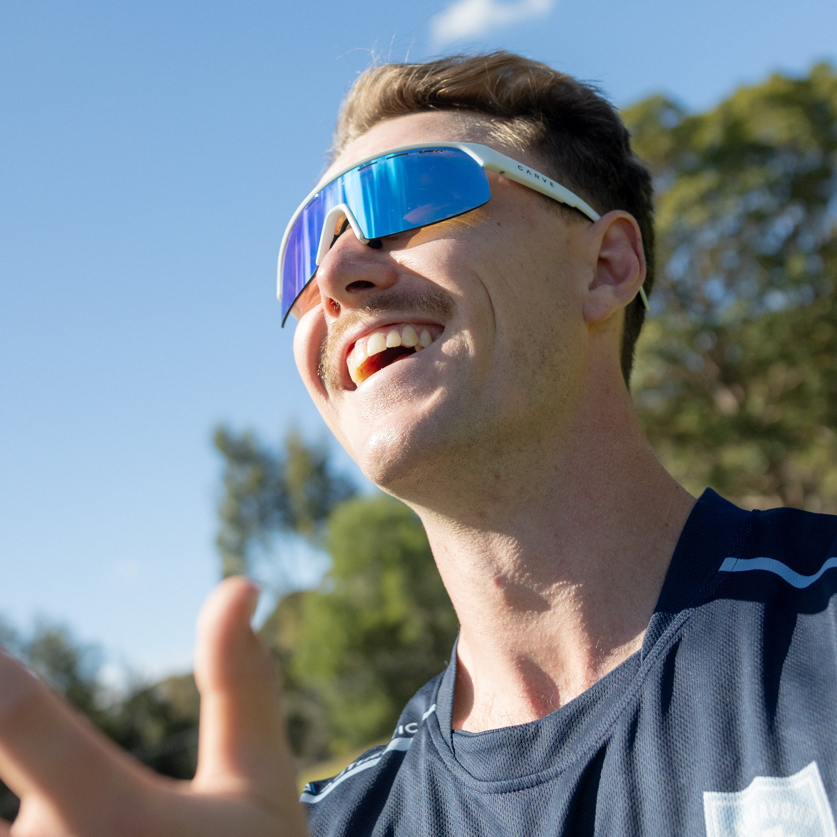 Person wearing reflective sunglasses outdoors with trees and blue sky in the background