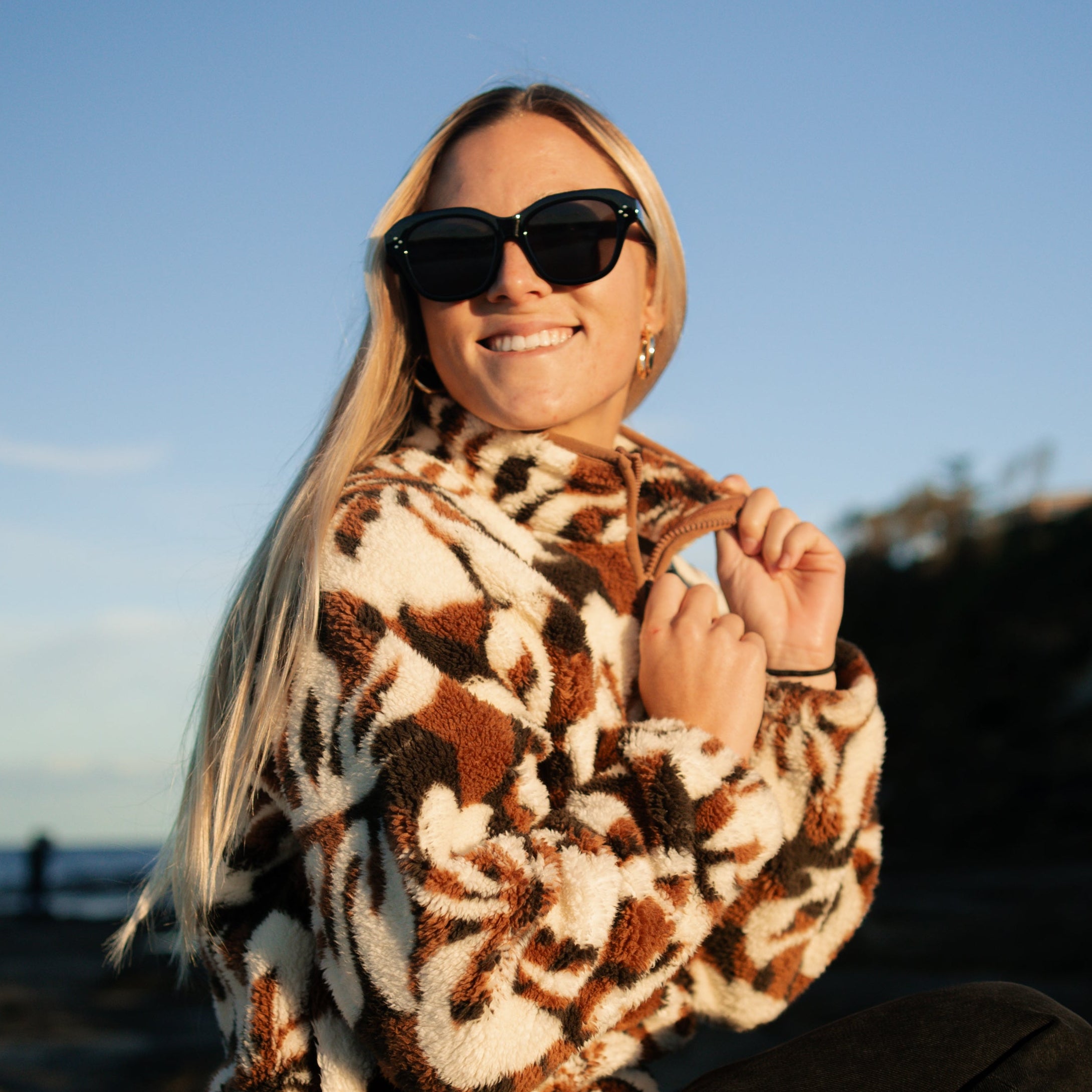 Person wearing sunglasses sitting on a rock by the ocean.