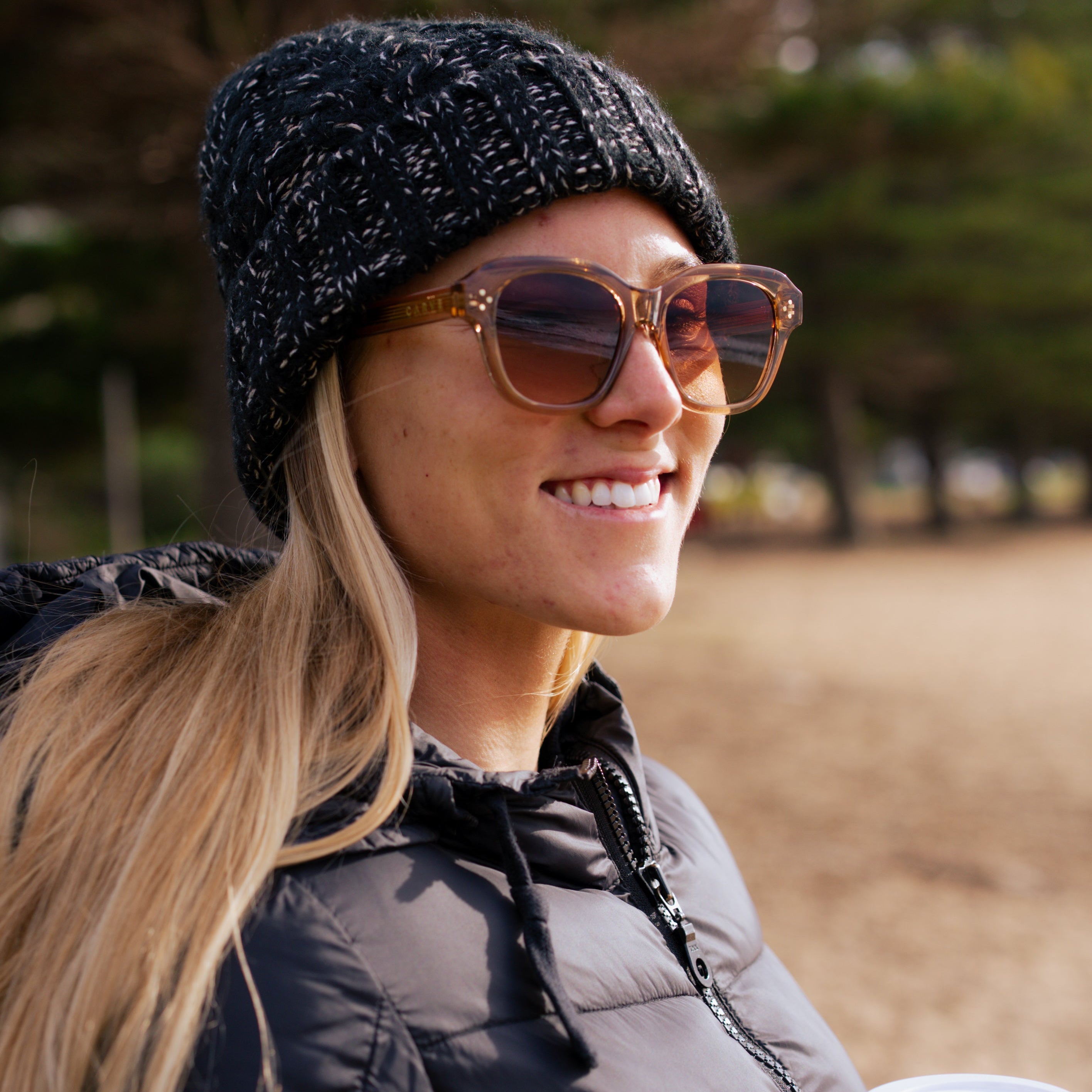 Woman wearing sunglasses and a black beanie, holding a coffee outdoors.