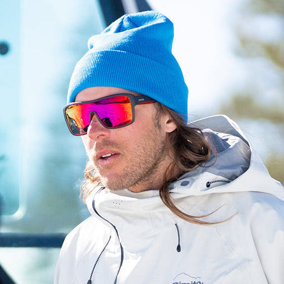 Person wearing a white jacket and blue beanie with sunglasses, standing in front of a vehicle.
