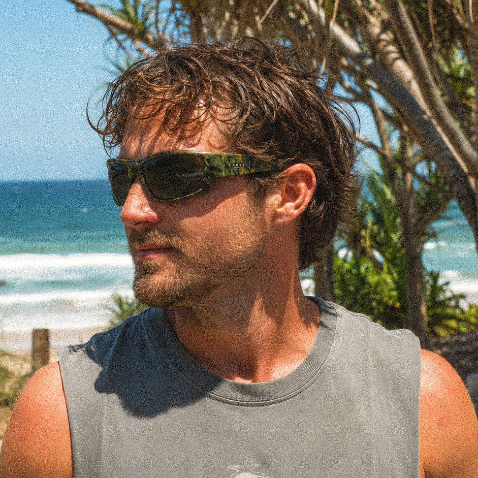 A man wearing dark sunglasses with a green and black patterned frame and a gray sleeveless shirt, standing outdoors on a beach path with trees and the ocean in the background.