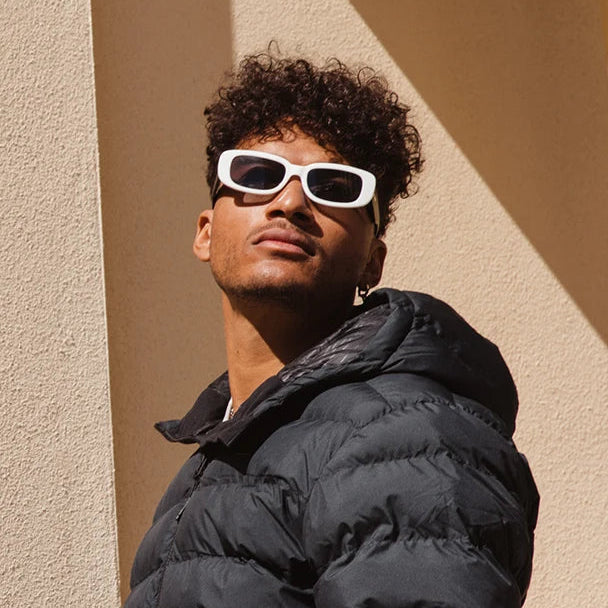 Man wearing a black puffer jacket and white sunglasses against a beige wall.