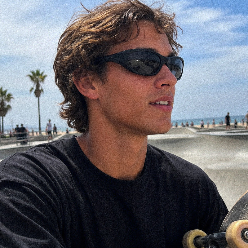 A man outdoors at a skatepark or beach area wearing black sunglasses and a black t-shirt, holding a skateboard. Palm trees, water, and a blue sky are visible in the background.