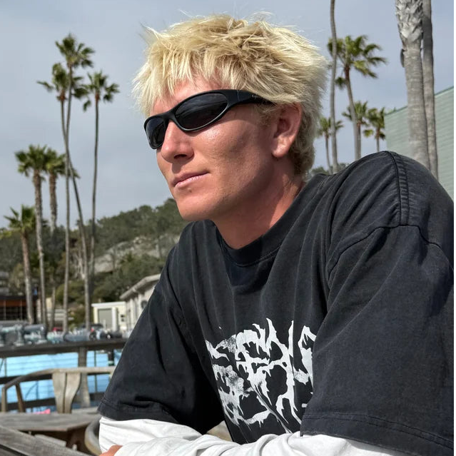 A man with blonde hair and sunglasses leans on a wooden railing outdoors, wearing a black graphic t-shirt over a white long-sleeve shirt, with palm trees and a cloudy sky in the background.